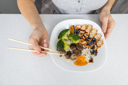 A girl in a gray shirt in the kitchen at home holds a plate of golden rice, shiitake mushrooms, chicken with unagi sauce and broccoli in her hands. The dish is decorated with black and white sesame seeds. Asian food concept.の写真素材