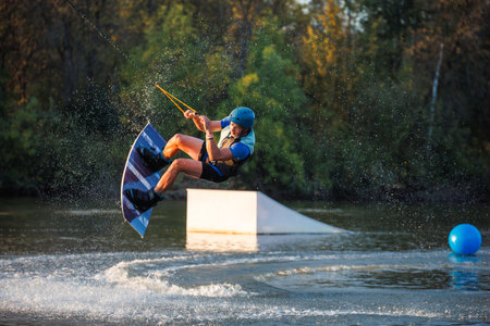 An athlete does a trick from a springboard. A rider jumps on a wakeboard against a background of a green forest. Sunset on the lake.の写真素材