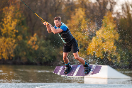 An athlete does a trick from a springboard. A rider jumps on a wakeboard against a background of a green forest. Sunset on the lake.の写真素材