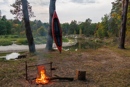 Camping on the bank of a forest river. The fire is burning. Red Kayak against a background of water and trees. Tourist parking in the evening.の写真素材