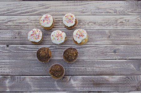 Tasty muffins with vanilla and chocolate cream decorated with sugar candy. Sweet pastries,  cakes on a wooden background in a shape of a heartの写真素材
