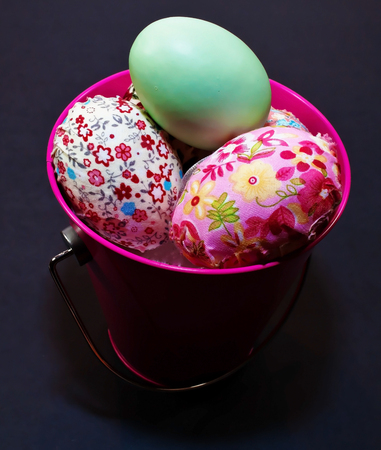 Several colorful Easter eggs in a pink bucket on a black background. The bucket is shown in faded light with the eggs illuminatedの写真素材