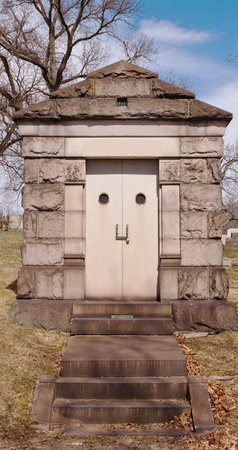 A mausoleum in a cemetery with bare winter trees behind it and bright blue and white clouds behind...