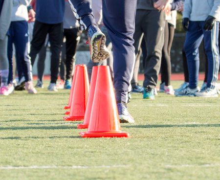 A runner does a dead leg drill over orange cones on a green turf field while his teammates watchの写真素材