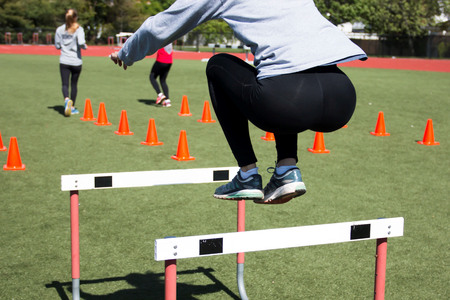 High school girls working out on a turf field by jumping over hurdles and doing running drills over orange cones.の写真素材