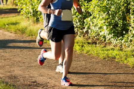 Two runners are racing a 5K in a New York State Park on a dirt path in the early evening ovee the...