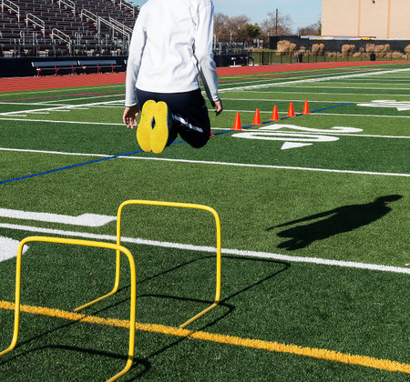 A high school track and field athlete jumps over yellow hurdles with orange cones in the background...