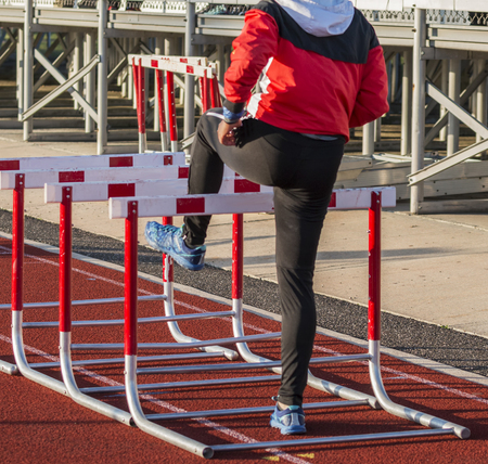 A high school girl track and field athlete is warming up for her hurdle race by walking over hurdles on a red track in her sweatsuit.の写真素材