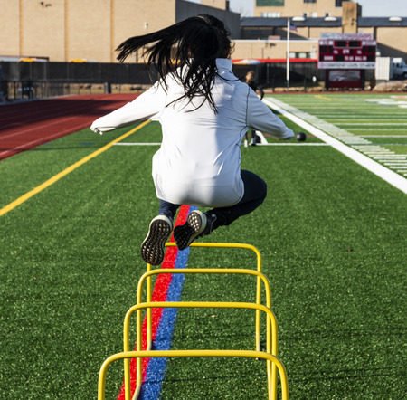 High school teenage girl jumping over two foot yellow hurldes that are set on a green turf field during track and field practice.の写真素材