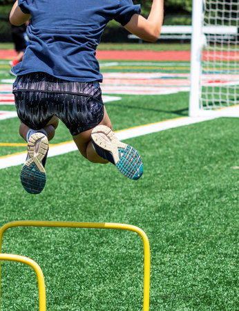 A high school female athlete is jumping over two feet yellow hurdles on a green turf field during...
