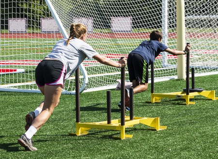 A high school girls and boy are pushing yellow weighted sleds on a green turf field during strength practice.のeditorial素材