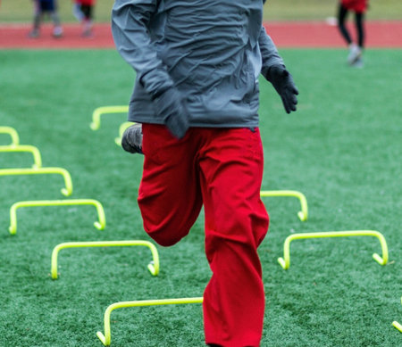 A high school runner is sprinting over yellow mini hurdels on a green turf field on a cold day in...