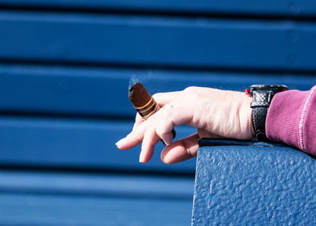 A man is resting his arm on a blue bench with the end of a cigar resting between his fingers with a...