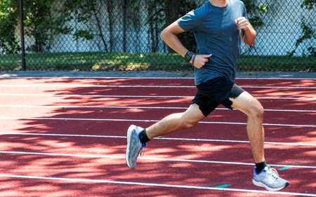 A high school track and field runner is running fast on a red track in the shade.