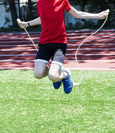 A young teenage boy in a red shirt is jumping rope on a green turf field with a red track in the backgound in sunshine.の写真素材