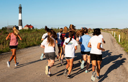 Fire Island, NEw York, USA - 29 August 2019: High school girls cross country team running with their coach passing the Fire Island Lighthouse on a sunny summer evening.のeditorial素材