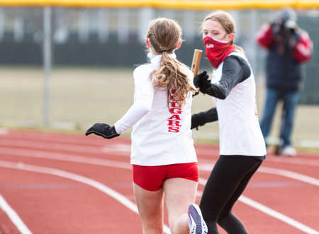 Hicksville, New York, USA - 17 January 2021: Girls exchanging the baton during a relay running race wearing face mask gaiters because of the coronavirus COVID-19 pandemic with selective focus blurring the background.のeditorial素材