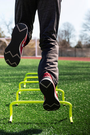 Rear view of a male athlete jumping on one leg over yellow mini hurdles on a green turf field during...