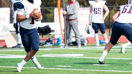 A high school football quarterback is dropping back during a during a passing play crossing his legs with both hands on the ball.の写真素材