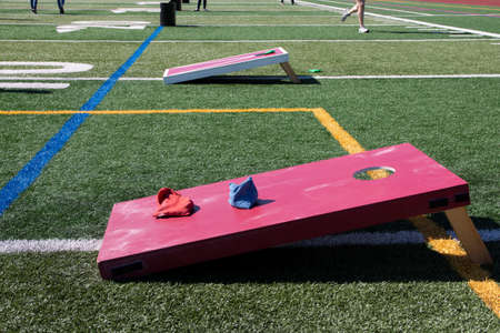 A high school turf field is set up with red and white wooden cornhole games and other backyard games for kids to play and have fun in the sun.の写真素材