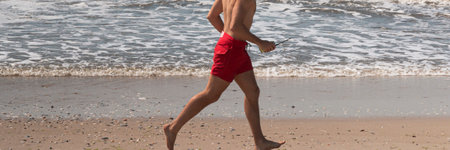 Side view of one male lifeguard running shirtless on the beach in red shorts close to the waterの写真素材