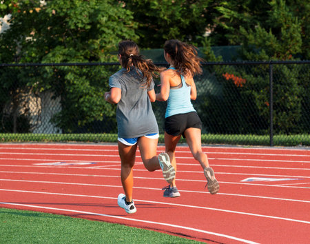 Rear view of two high school girls running together on the first turn of an outdoor track during summer camp track and field training.の写真素材