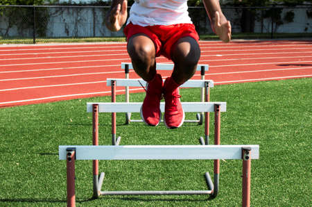 Front view of an African American high school boy jumping over track hurdles on a turf field during strength and agility practice.の写真素材