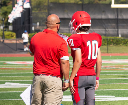 Rear view of a high school head football coach going over the next play with his quarterback on the...