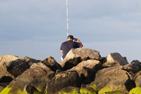 Close up of a man sitting on a rock jetty that has green moss at Sunken Meaow State Park fishing.の写真素材