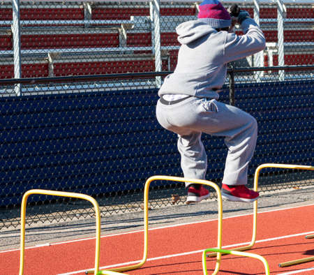A high school boy track and field athlete jumping over yellow hurdles on a winter mornings team practice wearing a sweatsuit, gloves and a hat to stay warm.の写真素材