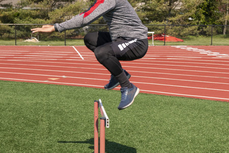 Side view of a male high school athlete jumping over a track hurdle on green truf close up.