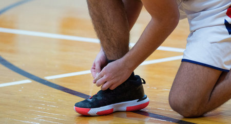 A high school basketball player is on his knee tying his shoes during a game indoors.