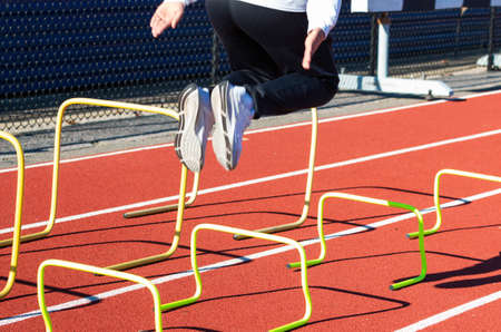 A high school track and field runner is jumping over yellow mini hutdles on a red track wearing white shoes.の写真素材