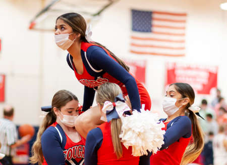 West Islip, New York, USA - 8 February 2022: High school cheerleaders buiding a pyramid while performing for the fans during a basketball game indoor wearing facemasks.のeditorial素材