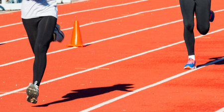 Two girls wearing black spandex while running fast on a red track past an orange cone.