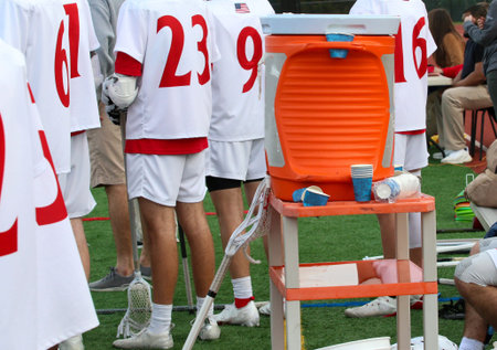 Orange water cooler on a stand by the athletes on the bench of a lacrosse game with paper cups.