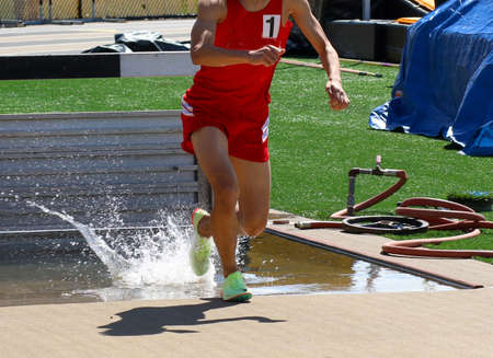 A high school runner wearing the number one on his red uniform exiting the steeplechase water pit with a little but of splashing during a race.の写真素材