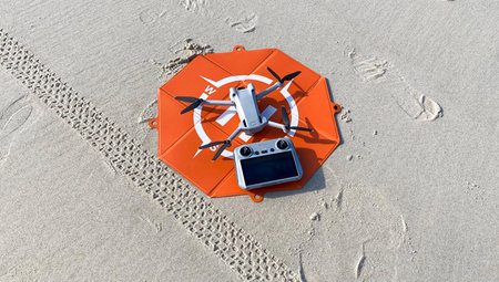 One drone with its controller on an orange takeoff and landing pad resting on the sand at a beach.