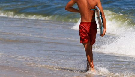 Rear view of a young man walking on the waters edge carrying a surfboard wearing a red bathingsuit at Gilgo Beach on Long Island.の写真素材