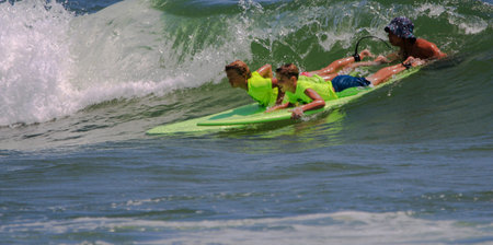 Gilgo Beach, New York, USA - 25 July 2023: Surf camp counselor holding two boys in neon green shirts...