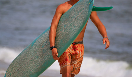 Surfer carrying his surboard on the shore of the beach with a blurred background.