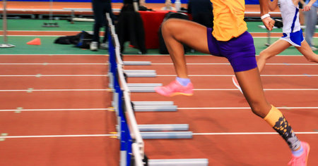 Hurdle runners going over hurdles in a race indoor during a track competition.