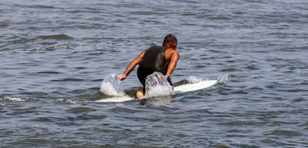 Rear view of a man kneeling on his surfboard paddling out to the waves at Gilgo Beach on Long...