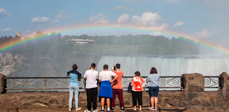 Niagara Falls, Onterio, Canada - 31 July 2023: Groups of people stand by a railing, watching a vivid rainbow arching over Niagara Falls under a clear sky.の写真素材