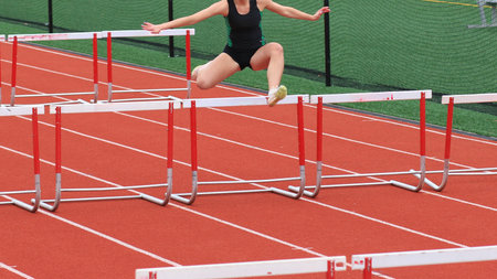 Female athlete jumps over hurdles on a vibrant red track, showcasing her speed and skill.の写真素材