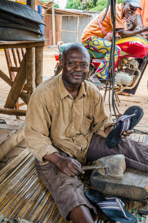 Abomey, Benin - March 07, 2014: Smiling African man sitting on ground and manufacturing tire flip flops in streetのeditorial素材