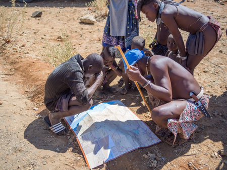 Opuwo, Namibia - July 25, 2015: Group of Himba men and boys leaning over and looking at map of...