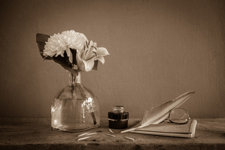 Still life with white dahlias in bottle, feather pen in ink bottle and old glass on brown book,の写真素材