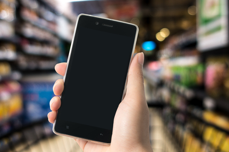 A man holding modern smart phone with blank screen in hand with supermarket or department store defocused background. Closeup shot.の写真素材