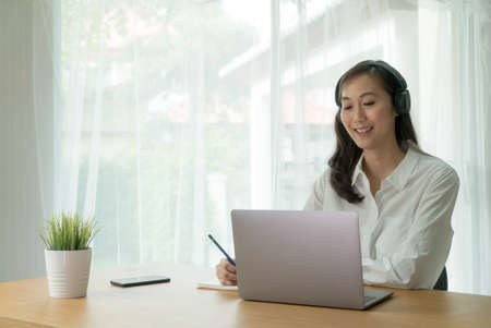 Asian female Smiling and video call online from laptop notebook computer waring wireless headphone and making notes at the desk.の写真素材